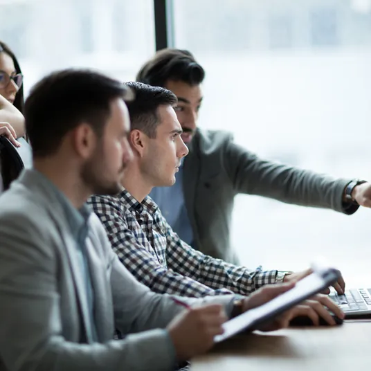 Un grupo de personas están sentadas en una mesa con un hombre señalando algo.